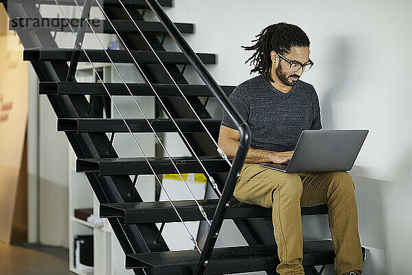 Selbstbewusster Geschäftsmann mit Dreadlocks benutzt Laptop-Computer  während er auf Stufen im Büro sitzt