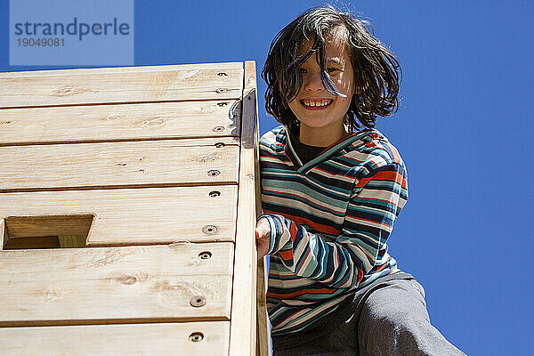 Ein glückliches Kind lehnt sich von einer hohen Holzwand vor blauem Himmel herab