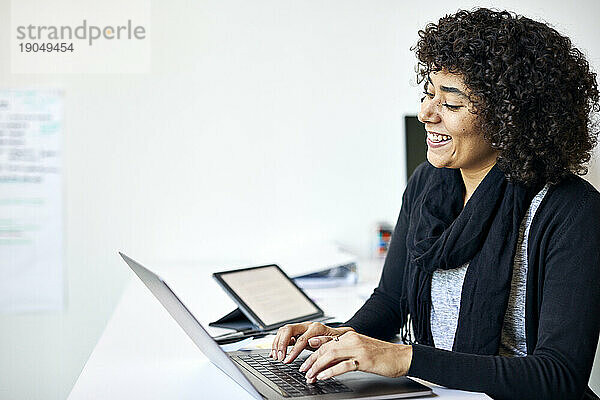 Lächelnde Geschäftsfrau mit lockigem Haar benutzt Laptop-Computer auf Schreibtisch an der Wand im Büro