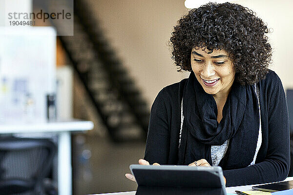 Lächelnde Geschäftsfrau mit lockigem Haar benutzt Tablet-Computer auf dem Schreibtisch im Büro