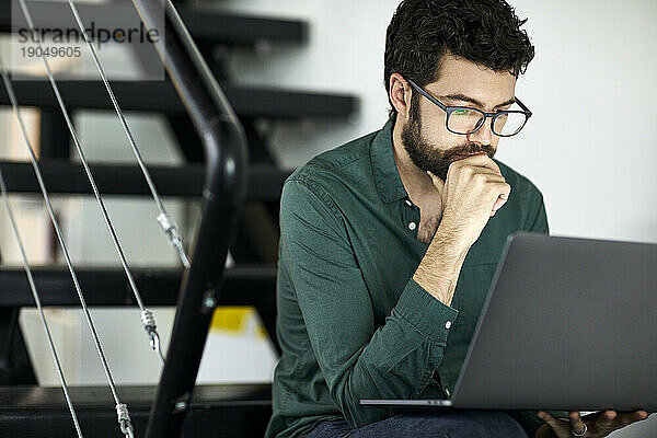 Selbstbewusster bärtiger Geschäftsmann benutzt Laptop-Computer  während er auf Stufen im Büro sitzt