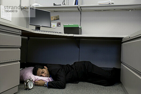 Ein Mann  der bei der Arbeit unter seinem Schreibtisch in seiner Kabine schläft.