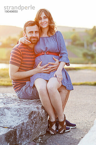 Mann und schwangere Frau im Kleid umarmen sich im Sommer bei Sonnenuntergang