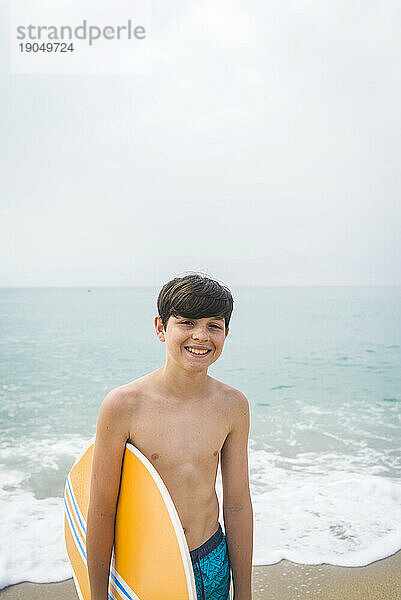 Vorderansicht eines jungen Teenagers  der am Strand steht und ein Surfbrett hält