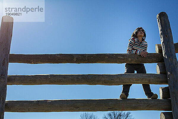 Ein nachdenkliches Kind steht auf einem hohen Holzzaun vor blauem Himmel