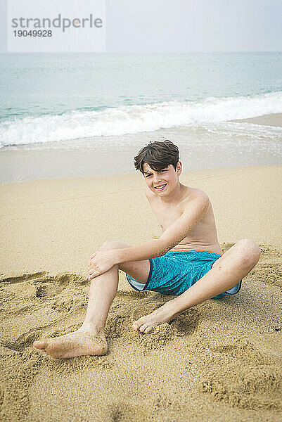 Lächelnder kleiner Junge in Badebekleidung sitzt am Strand und blickt in die Kamera