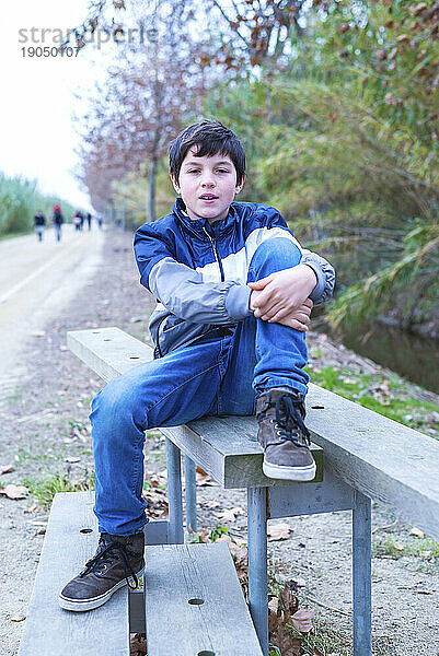 Ernsthafter junger Mann sitzt auf einem Holzgeländer und hat die Arme um die Beine geschlungen