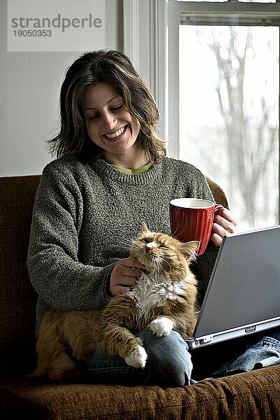 Frau entspannt sich mit ihrer Katze auf einem Stuhl  während sie an einem Laptop arbeitet.