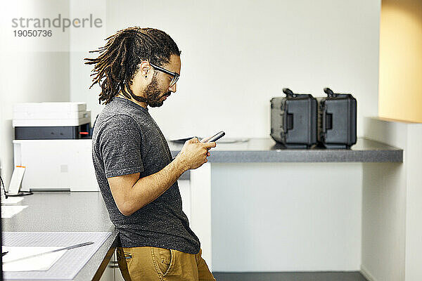 Seitenansicht eines selbstbewussten Geschäftsmannes  der sein Smartphone nutzt  während er im Büro am Tisch steht