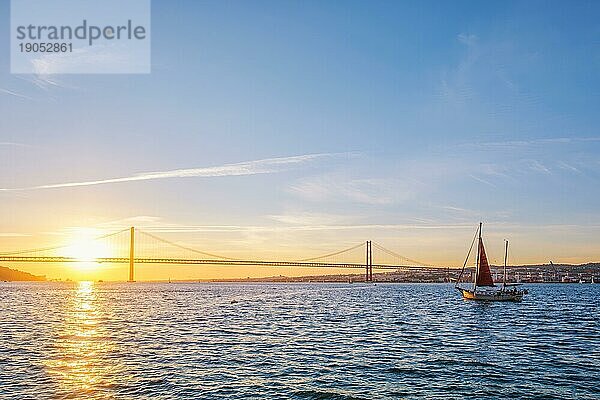 Blick auf die Brücke 25 de Abril  berühmtes touristisches Wahrzeichen von Lissabon  die Lisboa und Almada über den Fluss Tejo verbindet  mit der Silhouette einer Touristenyacht bei Sonnenuntergang. Lissabon  Portugal  Europa
