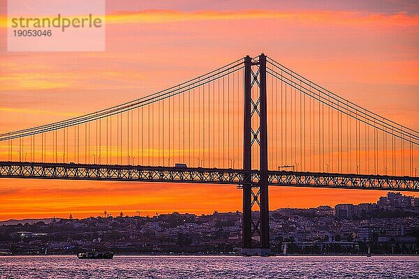 Blick auf die Brücke 25 de Abril  berühmtes touristisches Wahrzeichen von Lissabon  die Lisboa und Almada über den Fluss Tejo verbindet  mit der Silhouette eines Touristenboots bei Sonnenuntergang. Lissabon  Portugal  Europa