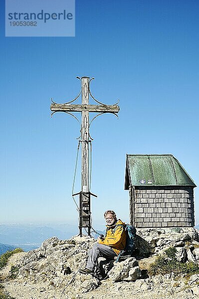 Wanderer  Senior  65  rastet auf dem Geigelstein neben Gipfelkreuz und Kapelle  Sachrang  Schleching  Chiemgau  Oberbayern  Deutschland  Europa