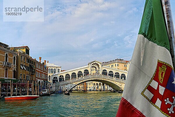 Schöner Blick auf die italienische Seeflagge und die Rialtobrücke oder Ponte Rialto am Canal Grande in Venedig  Italien. Gondel Service Station auf der linken Seite. UNESCO Weltkulturerbe Stadt. Tageslicht  Sonnenschein  weiche Wolken. Selektiver Fokus