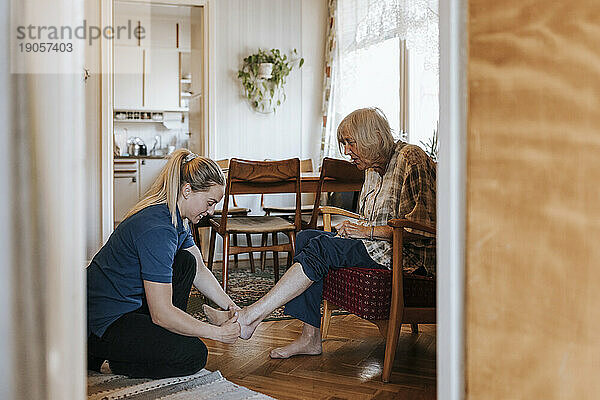 Seitenansicht einer Pflegehelferin  die einer älteren Frau beim Tragen von Socken hilft  während sie zu Hause sitzt