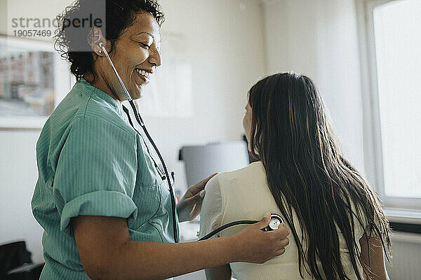 Seitenansicht einer lächelnden reifen Ärztin  die eine junge Frau mit Stethoskop in einer Klinik untersucht