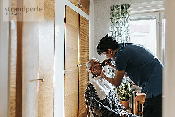Seitenansicht eines männlichen Pflegehelfers  der den Schnurrbart eines älteren Mannes zu Hause trimmt