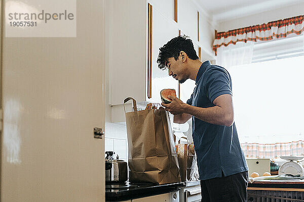Seitenansicht eines männlichen Pflegehelfers beim Auspacken von Lebensmitteln aus einer Tüte in der Küche zu Hause
