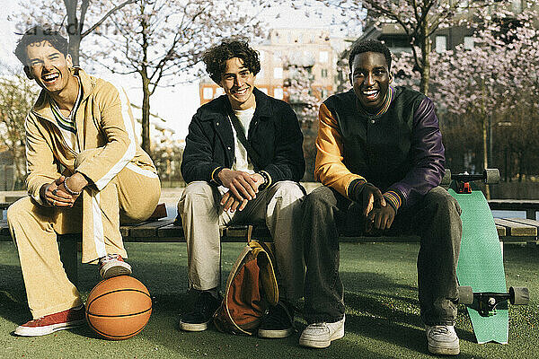 Glückliche multirassische männliche Freunde  die lachend auf einer Bank sitzen  während eines sonnigen Tages