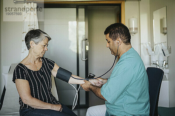 Männlicher Arzt  der im Krankenhaus sitzend den Blutdruck einer älteren Frau kontrolliert