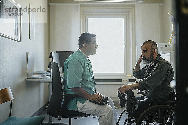 Seitenansicht eines reifen männlichen Arztes  der mit einem im Rollstuhl sitzenden Patienten in einer Klinik spricht