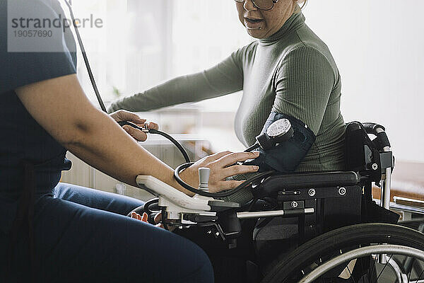 Mittelteil einer Krankenschwester  die den Blutdruck einer im Rollstuhl sitzenden Frau in einer Klinik überprüft
