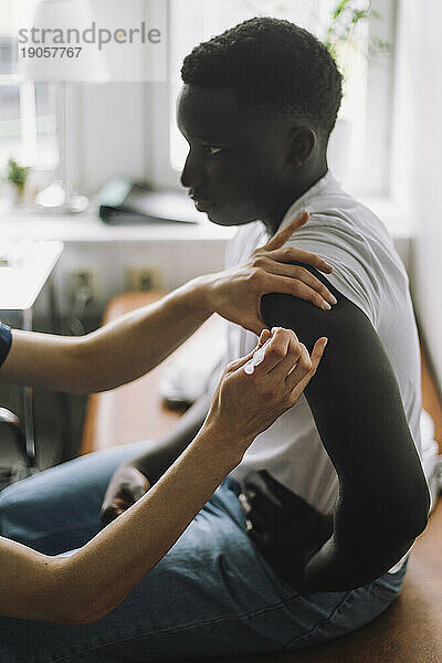 Seitenansicht eines männlichen jugendlichen Patienten  der sich in der Klinik sitzend am Arm impfen lässt