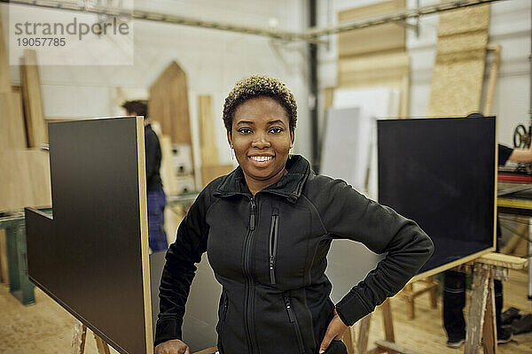 Porträt einer jungen Schreinerin  die in der Werkstatt steht und die Hand auf die Hüfte legt