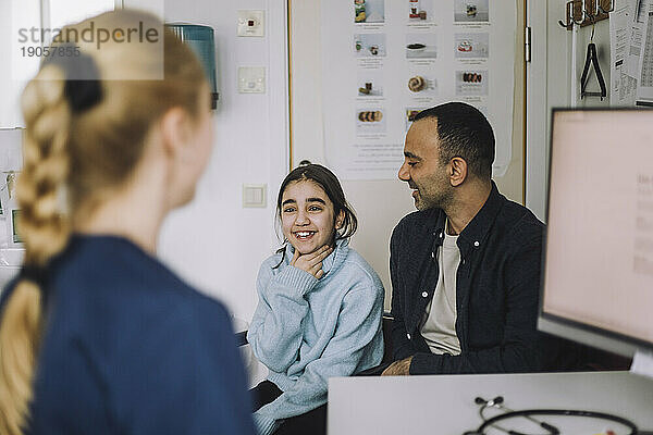 Lächelndes Mädchen erklärt dem Arzt  während es mit seinem Vater bei einem Besuch in der Klinik sitzt