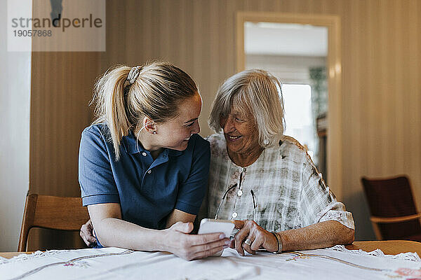 Lächelnde weibliche Betreuungsassistentin  die ihr Smartphone mit einer älteren Frau teilt  während sie am Esstisch im Haus sitzt