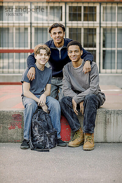 Porträt eines lächelnden Teenagers mit Armen um männliche Freunde  die auf dem Beton eines High-School-Campus sitzen