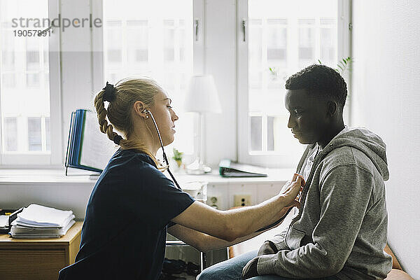 Seitenansicht einer Mitarbeiterin im Gesundheitswesen  die den Herzschlag eines männlichen Patienten im Teenageralter in einer Klinik überprüft