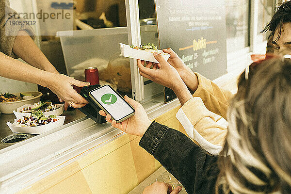 Frau  die per Tap-to-Pay-Methode mit ihrem Smartphone bezahlt  während sie Essen am Kiosk kauft