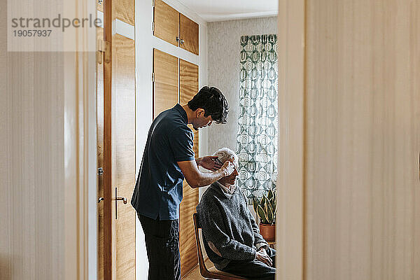 Seitenansicht eines männlichen Pflegehelfers  der die Haare eines älteren Mannes kämmt  der zu Hause sitzt