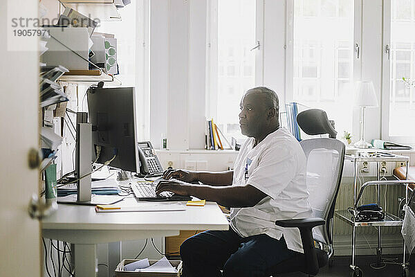 Reifer männlicher Arzt arbeitet am Computer  während er am Schreibtisch in der Klinik sitzt