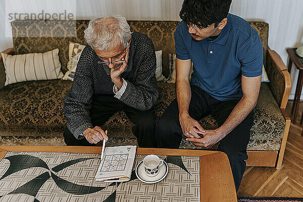 Hochformatige Ansicht eines älteren Mannes  der mit einem männlichen Betreuer zu Hause sitzt und ein Soduko-Puzzle spielt