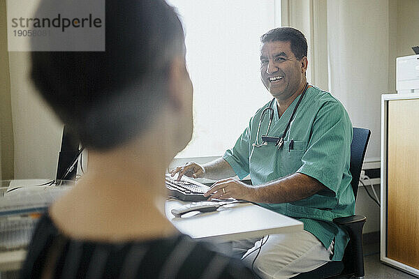 Glücklicher männlicher Arzt  der einen Patienten ansieht  während er einen Computer in einer Klinik benutzt