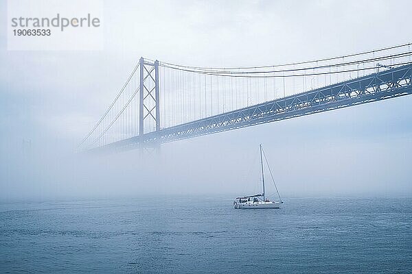 Blick auf die Brücke 25 de Abril  das berühmte touristische Wahrzeichen Lissabons  die Lisboa und Almada verbindet  im dichten Nebel mit darunter durchfahrenden Booten. Lissabon  Portugal  Europa