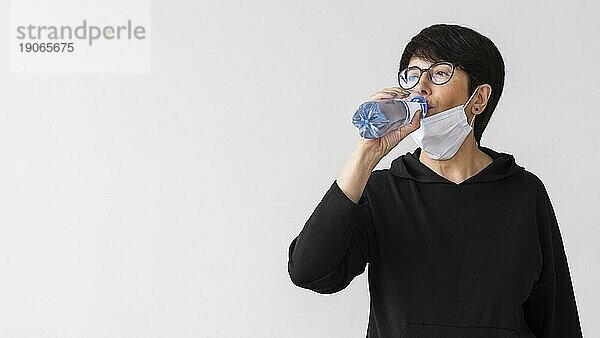 Frau mit medizinischer Maske trinkt Wasser aus einer Flasche