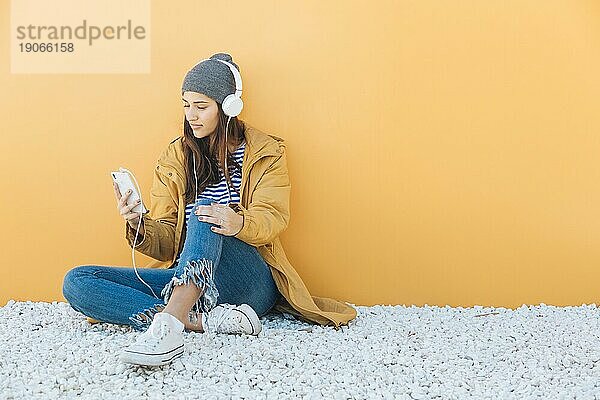 Frau  die auf einem Teppich sitzt und mit ihrem Smartphone Musik über Kopfhörer hört