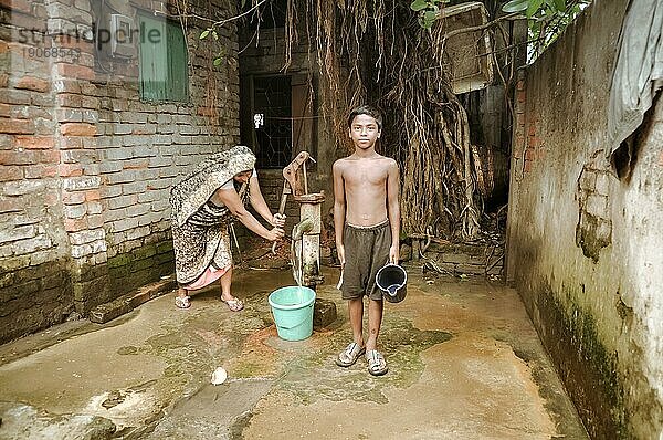 Khulna  Bangladesch  etwa im Juli 2012: Ein halbnackter Junge hält einen blaün Eimer in seiner Hand im Slum von Khula  Bangladesch. Seine Mutter pumpt Wasser aus einem Brunnen in den Eimer. Dokumentarischer Leitartikel  Asien