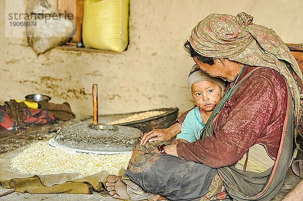 Dolpo  Nepal  ca. Mai 2012: Einheimische Frau mit Kopftuch sitzt mit einem Kind auf dem Boden in der Nähe einer steinernen Handgetreidemühle in Dolpo  Nepal. Dokumentarischer Leitartikel  Asien