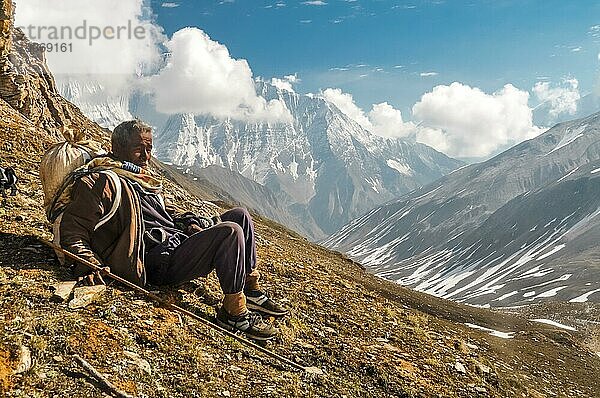 Dolpo  Nepal  ca. Juni 2012: Ein Einheimischer mit einer Tasche auf dem Rücken und einem Holzstock in der Hand sitzt auf dem Boden und ruht sich in den wunderschönen verschneiten Bergen in Dolpo  aus. Dokumentarischer Leitartikel  Asien