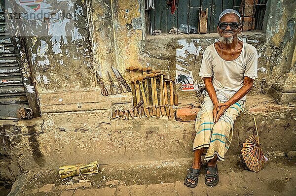 Dhaka  Bangladesch  etwa im Juli 2012: Alter lächelnder Mann mit weißem Bart sitzt auf einer Steinbank und verkauft Hämmer und Scheren auf der Straße in Dhaka  Bangladesch. Dokumentarischer Leitartikel  Asien