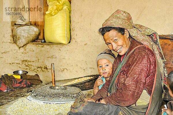 Dolpo  Nepal  ca. Mai 2012: Lächelnde Frau mit Kopfbedeckung und Kind sitzt auf dem Boden in der Nähe einer steinernen Handgetreidemühle in Dolpo  Nepal. Dokumentarischer Leitartikel  Asien