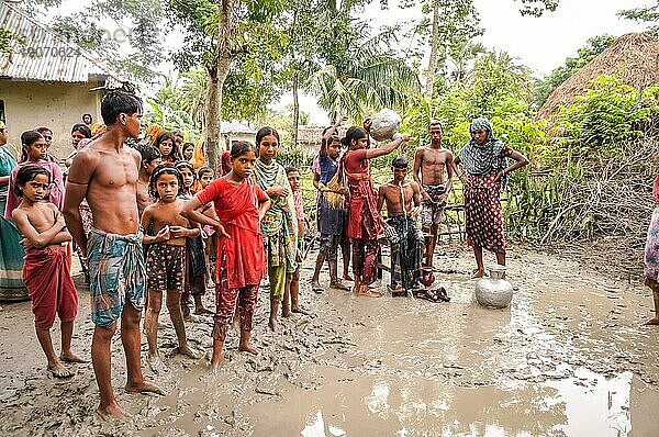 Paigacha  Bangladesch  ca. Juli 2012: Einheimische mit Kindern stehen im Schlamm und ein Mädchen in Rot gießt Wasser aus einem Krug auf den Kopf eines halbnackt sitzenden Jungen in Paigacha  Bangladesch. Dokumentarischer Leitartikel  Asien