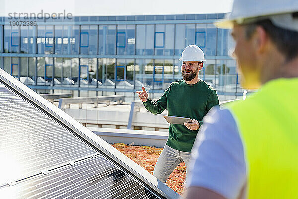 Mann mit Tablet-PC im Gespräch mit Kollegen auf dem Dach eines Firmengebäudes mit Solaranlagen