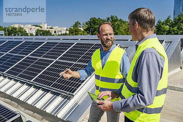 Zwei Männer mit Tablet-PC und reflektierenden Westen unterhalten sich auf dem Dach eines Firmengebäudes mit Solarpaneelen