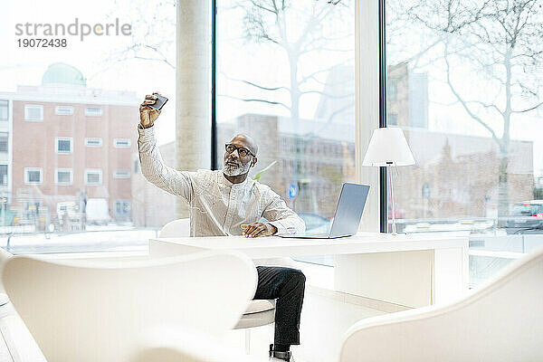 Porträt eines reifen Mannes mit Laptop  der am Schreibtisch sitzt und ein Selfie mit dem Smartphone macht