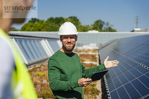 Mann mit Tablet-PC im Gespräch mit Kollegen auf dem Dach eines Firmengebäudes mit Solaranlagen