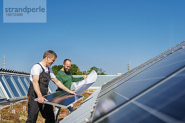 Handwerker installiert Solarpanel auf dem Dach eines Firmengebäudes  zusammen mit einem Kollegen  der den Plan hält